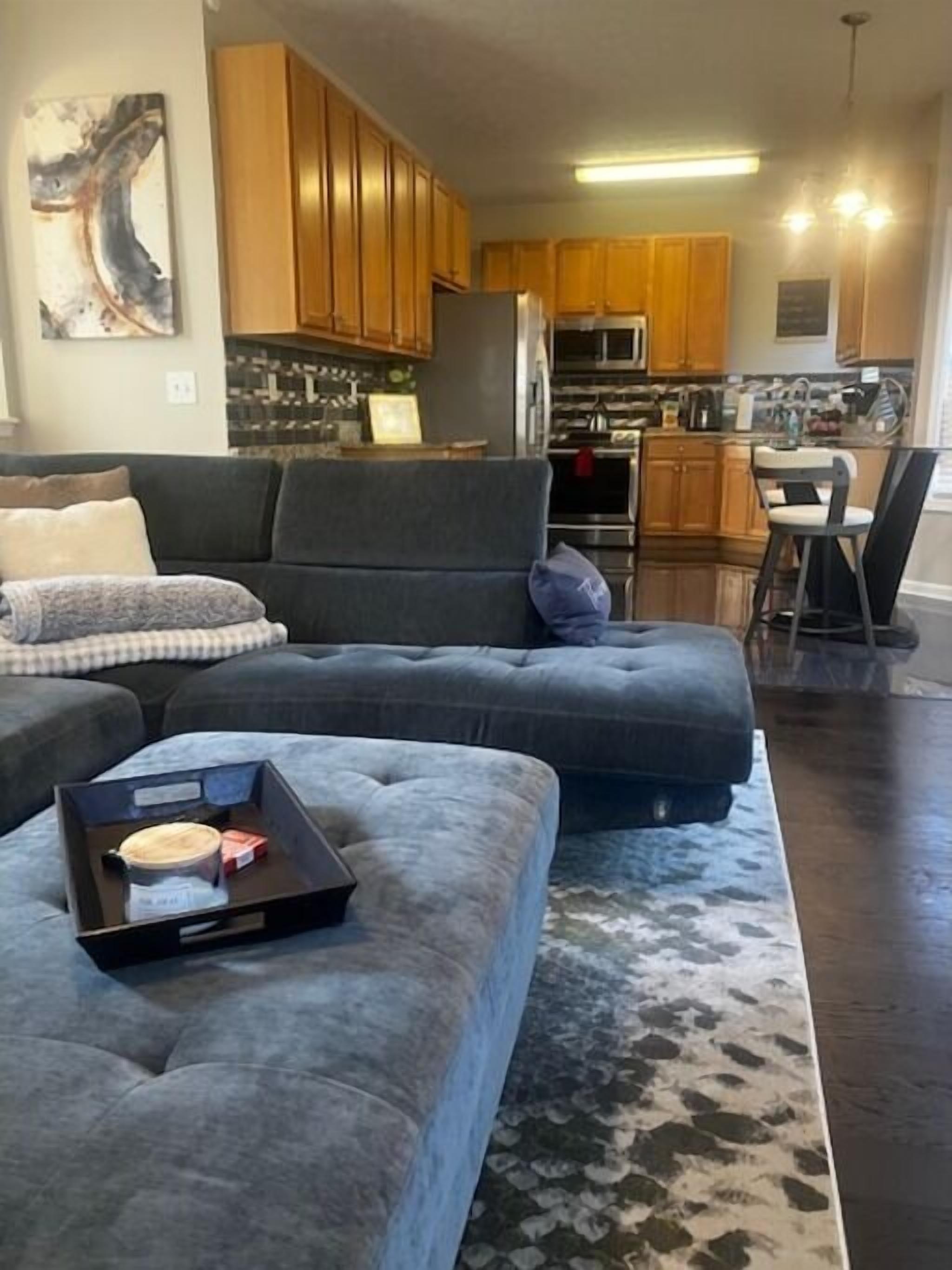 A modern living room with a grey sectional sofa, a dining area with a small table, and a kitchen with wooden cabinets and stainless steel appliances.