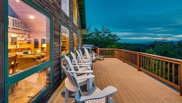 A wooden deck with white chairs, a wooden railing, and a view of a forested landscape.