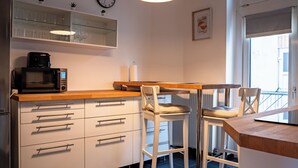 Eine moderne Küche mit weißen Schränken, einer Mikrowelle und einer Holz Arbeitsplatte. Zwei weiße Stühle und ein kleiner Tisch sind vorhanden. An der Wand befinden sich eine Uhr und ein gerahmtes Bild.