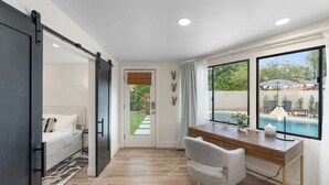Ein modernes Schlafzimmer mit einer Schiebetür, die auf einen Balkon führt, einem Schreibtisch mit einer Lampe und Blick auf einen Swimmingpool.