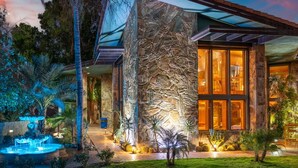 A modern house with a stone facade, large windows, and a well-lit garden with a fountain.
