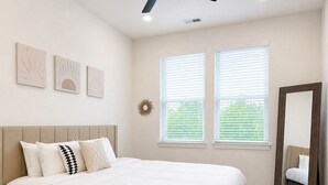 Un dormitorio con una cama, un ventilador de techo, dos ventanas con persianas y un espejo.