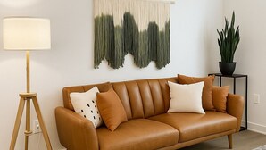 A brown leather sofa with cushions, a floor lamp, a macrame wall hanging, a potted plant, and a rug. - WorkerFriendly,Wash/Dry,WiFi, Pool,A+Location,Gym (Sherman)