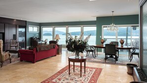 A spacious living room with a red sofa, a round table with a vase of flowers, and a view of a lake through large windows.