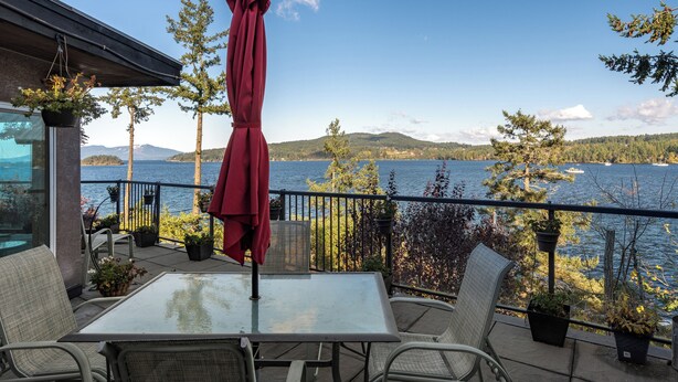 A balcony with a table and chairs, overlooking a lake with trees and mountains in the background.