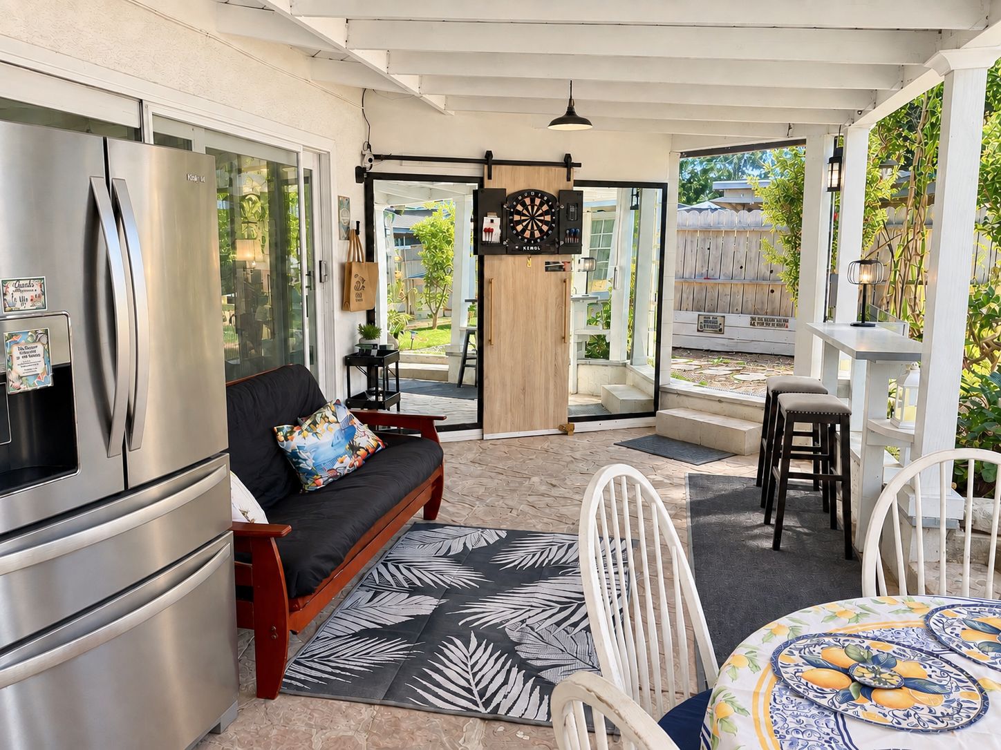 A modern kitchen with a stainless steel refrigerator, a black sofa with a colorful cushion, and a dining area with white chairs and a patterned tablecloth.