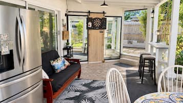 A modern kitchen with a stainless steel refrigerator, a black sofa with a colorful cushion, and a dining area with white chairs and a patterned tablecloth.
