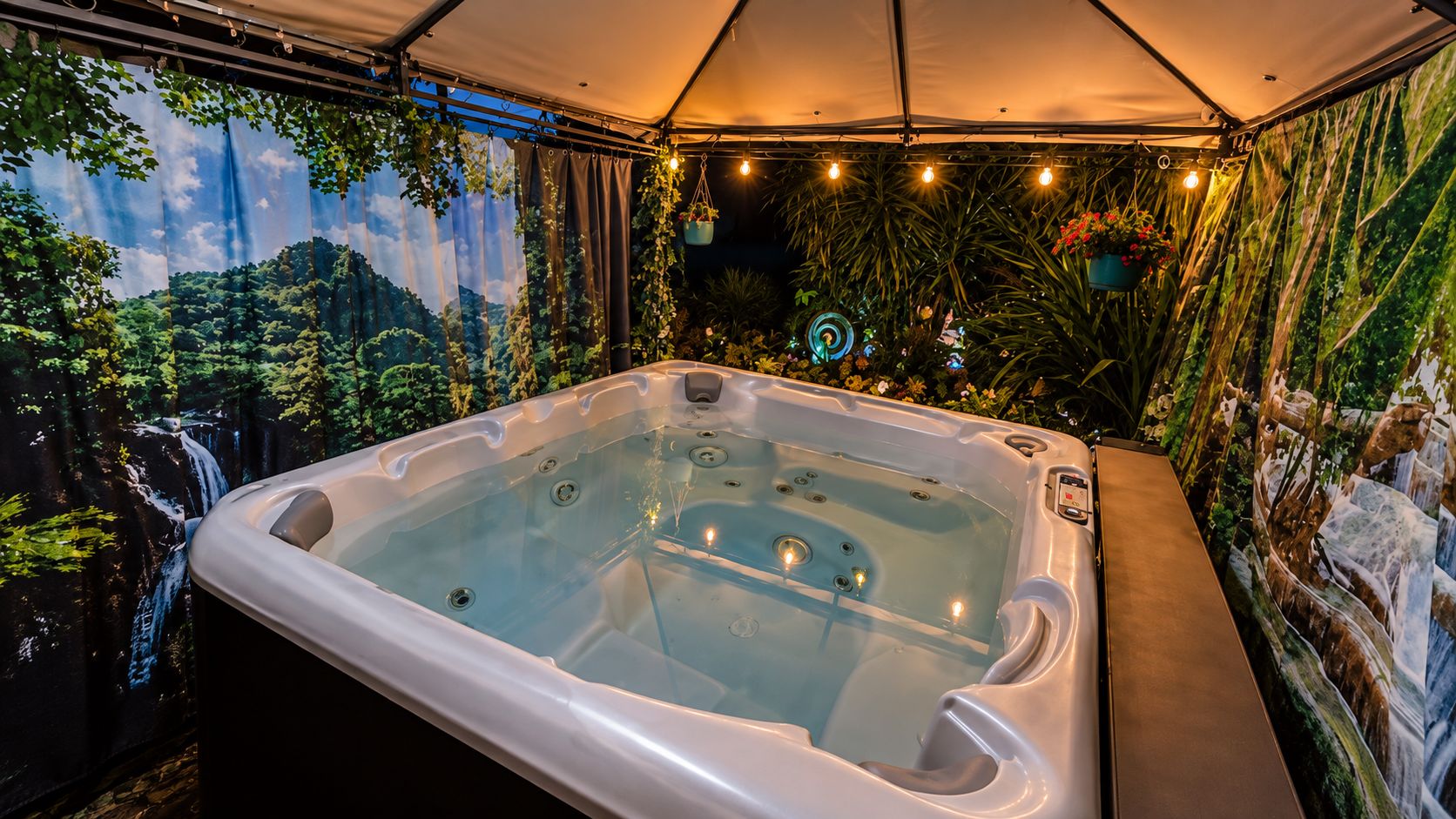 A hot tub in a tented area with a forest mural on the walls and hanging plants.