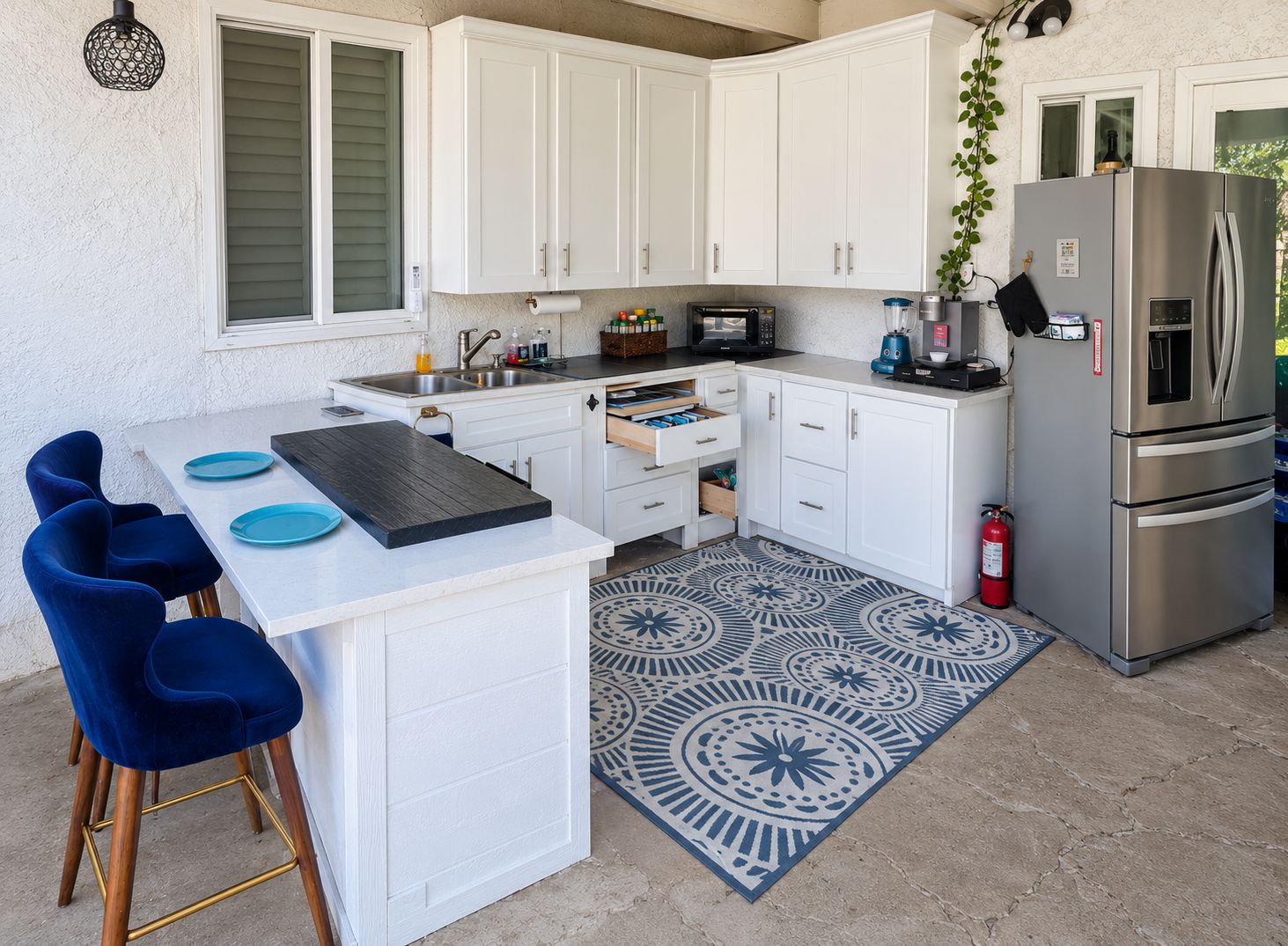 A compact kitchen with white cabinets, a stainless steel refrigerator, a microwave, and a sink. There are blue chairs and a patterned rug on the floor.