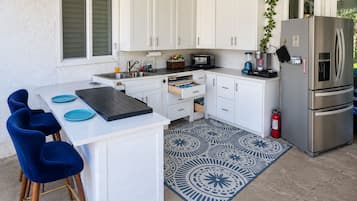 A compact kitchen with white cabinets, a stainless steel refrigerator, a microwave, and a sink. There are blue chairs and a patterned rug on the floor.