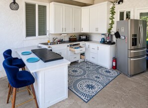 Une cuisine compacte avec des meubles blancs, un réfrigérateur en acier inoxydable, un four à micro-ondes et un évier. On y trouve des chaises bleues et un tapis à motifs sur le sol.