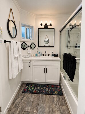 Une salle de bain avec un meuble-lavabo blanc, un miroir, une fenêtre, une douche et un tapis décoratif.