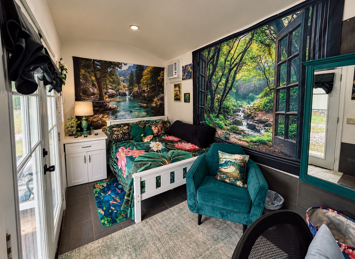 A bedroom with a large window, a bed with floral bedding, a teal armchair, and a view of a forest scene.