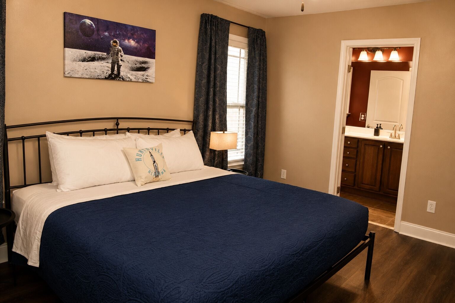 A bedroom with a large bed, a nightstand, a window with curtains, a bathroom visible through an open door, and a wall-mounted painting of an astronaut on the moon.