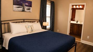 A bedroom with a large bed, a nightstand, a window with curtains, a bathroom visible through an open door, and a wall-mounted painting of an astronaut on the moon.