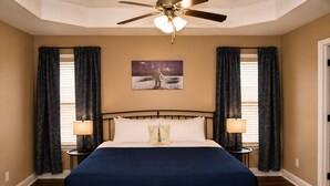 A bedroom with a bed, two bedside lamps, a ceiling fan, and a wall-mounted painting.