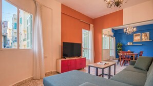A living room with a television, red cabinet, blue sofa, and a dining area with chairs and a table. - Muse 2-BR Apt in Mar Mikhael (Beirut)