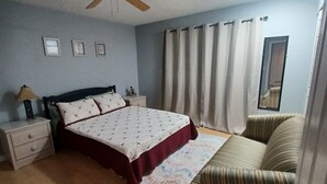 Une chambre à coucher comprenant un lit, une table de chevet, une chaise, un ventilateur de plafond et une fenêtre avec des rideaux.