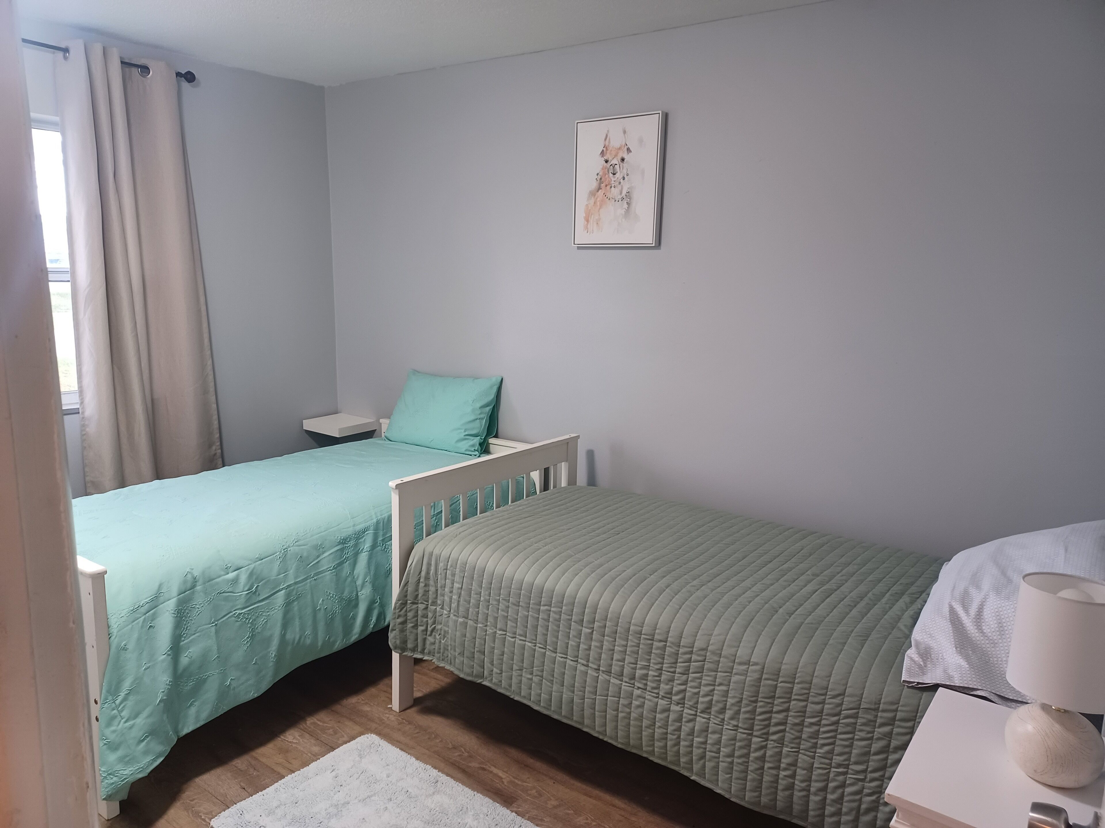 A bedroom with two single beds, a bedside table with a lamp, a framed picture on the wall, and a window with curtains.