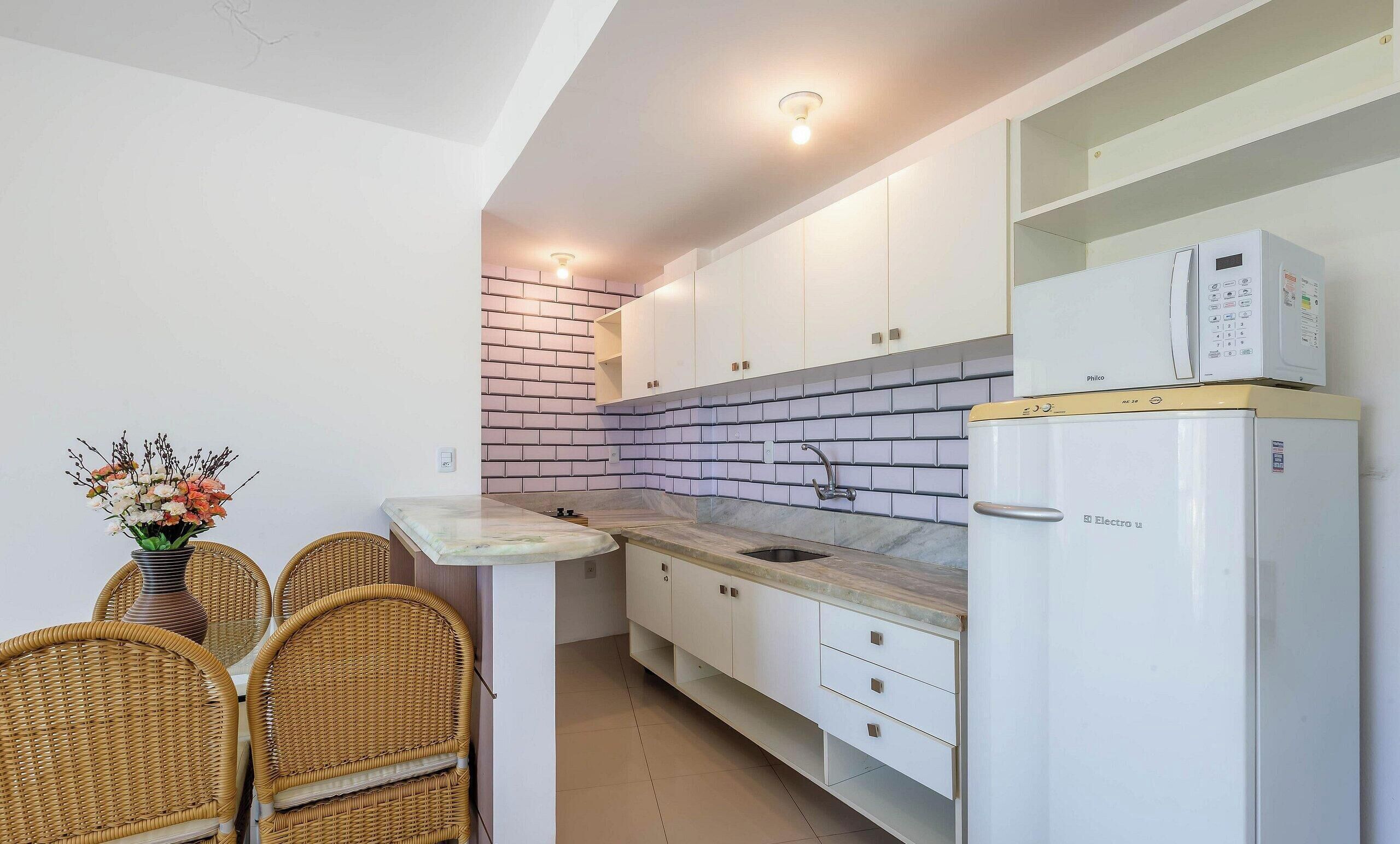 Una cocina compacta con armarios blancos, un salpicadero de ladrillo integrado, un refrigerador blanco y un comedor con sillas de mimbre y un jarrón con flores.