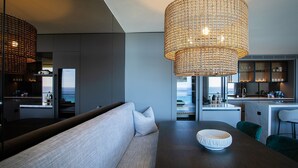 A modern kitchen with a large island, a dining area with a wooden table and a bowl, and a seating area with a sofa and cushions. - Sky Penthouse, Tamarama (Tamarama)