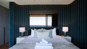 A modern hotel room with a large bed, two bedside lamps, a view of the sea through a window, and dark blue curtains. - Sky Penthouse, Tamarama (Tamarama)