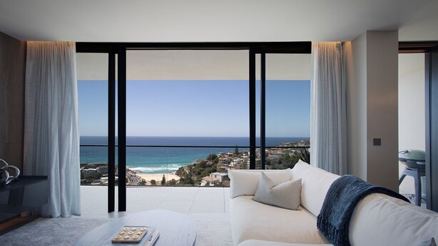 A modern living room with a large window overlooking a beach and ocean. - Sky Penthouse, Tamarama (Tamarama)