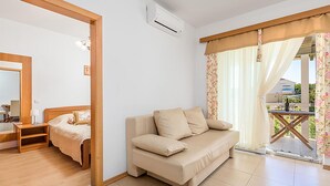 Moderni hotellihuone, jossa on beigenvärinen sohva, puinen ovi, pieni pöytä lampun kanssa ja näkymä ulos lasiovesta.