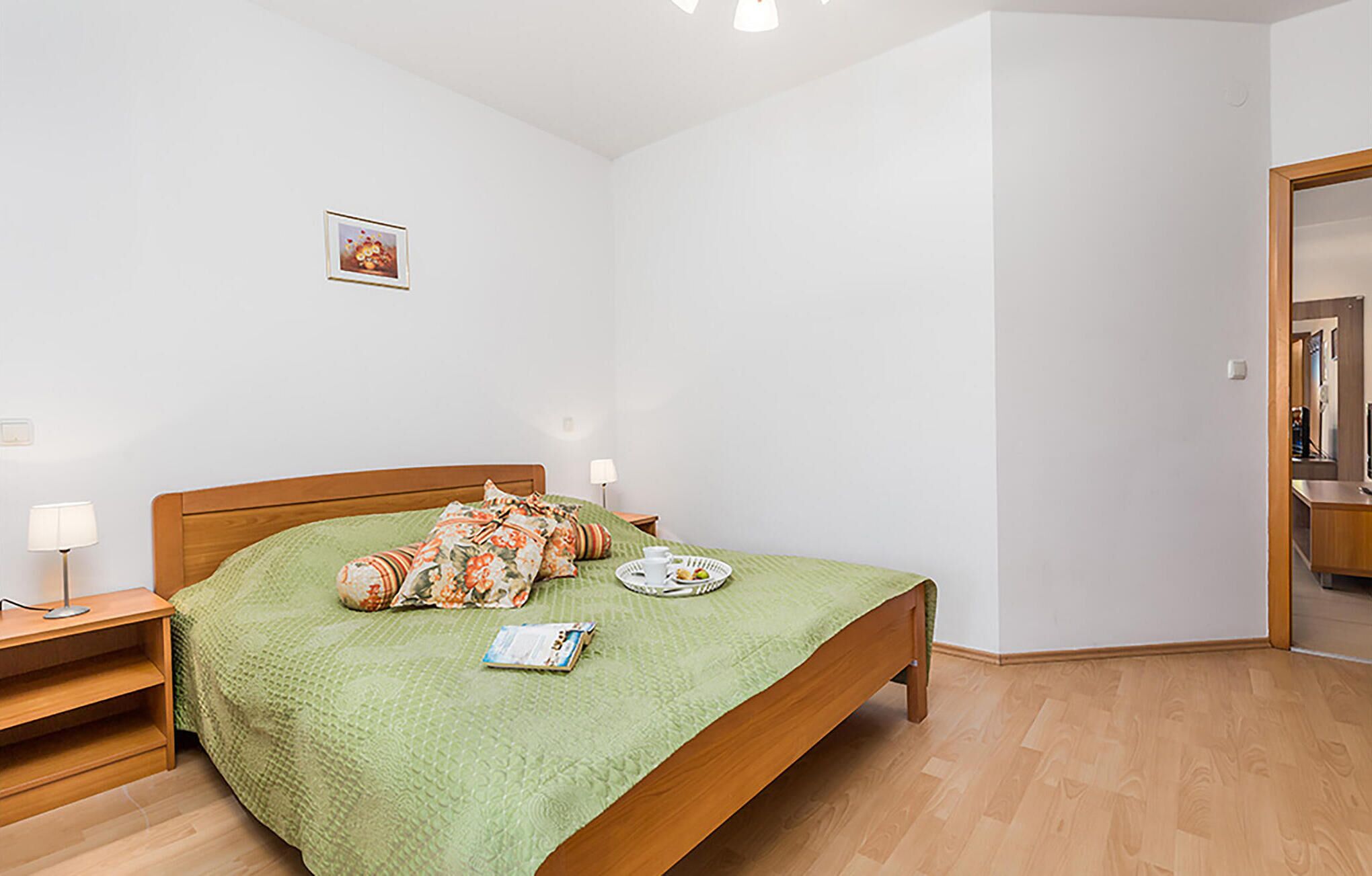 Un dormitorio ordenado con cama de madera, ropa de cama verde, una mesita de noche con lámpara y un cuadro pequeño en la pared.