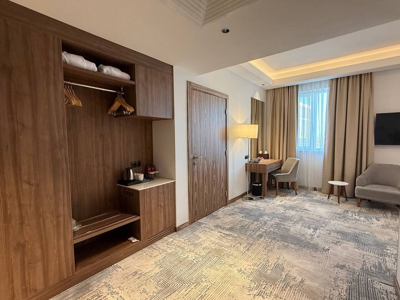 A hotel room with a wooden wardrobe, a desk with a lamp, a chair, and a sofa.