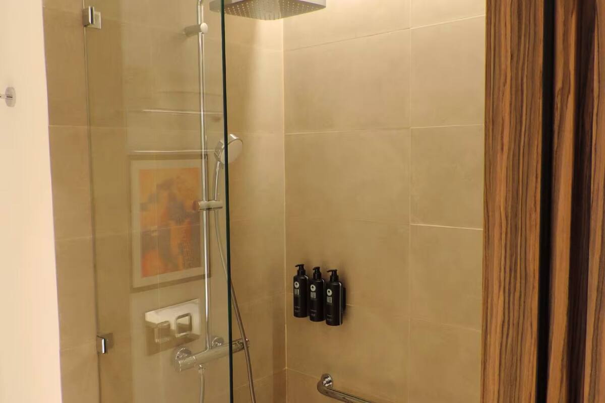A glass-enclosed shower with a handheld showerhead, a towel rack, and three toiletry bottles on the wall.