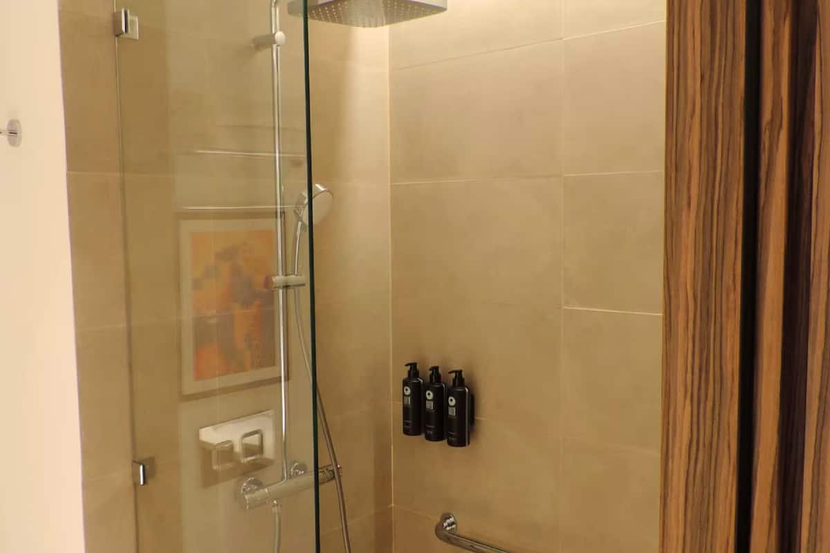 Une douche fermée par des parois en verre, dotée d’un pommeau de douche à main, d’un porte-serviettes et de trois flacons de produits de toilette fixés au mur.