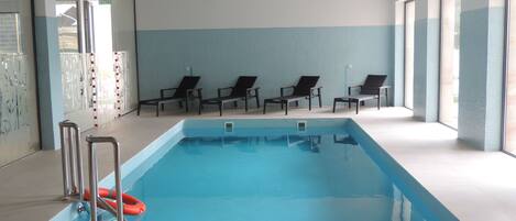 Una piscina interior con agua azul cristalina, rodeada de sillones de descanso negros y una pared con azulejos.