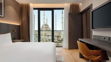 A hotel room with a large bed, a desk, a chair, a TV, and a view of a mosque with minarets.