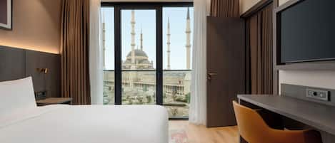 Et hotelværelse med en stor seng, et skrivebord, en stol, et fjernsyn og udsigt til en moské med minareter.