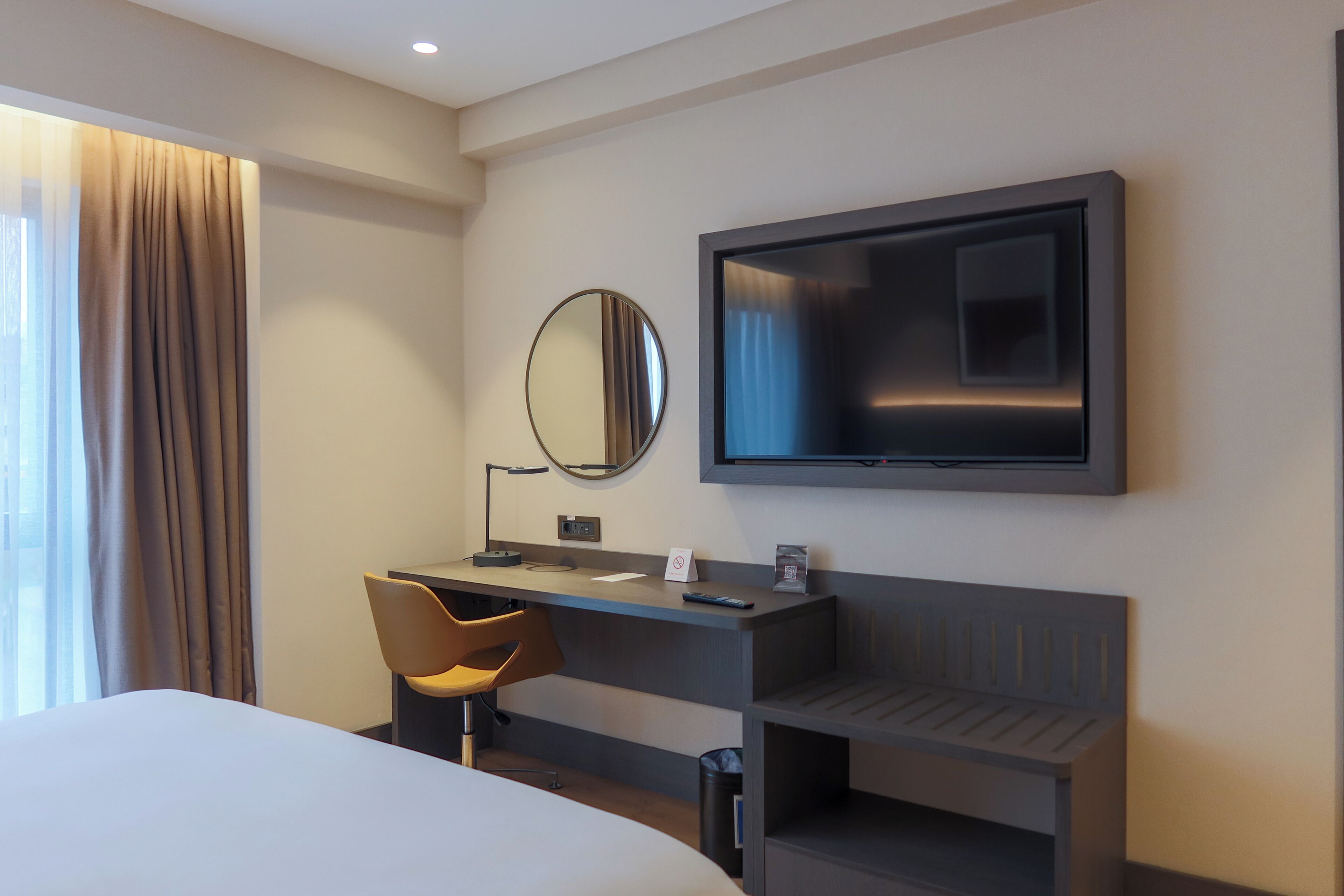 Et moderne hotelværelse med en seng, et skrivebord med stol, et fladskærms-tv og et stort spejl.