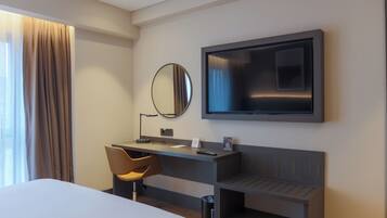 A modern hotel room with a bed, a desk with a chair, a flat-screen TV, and a large mirror.