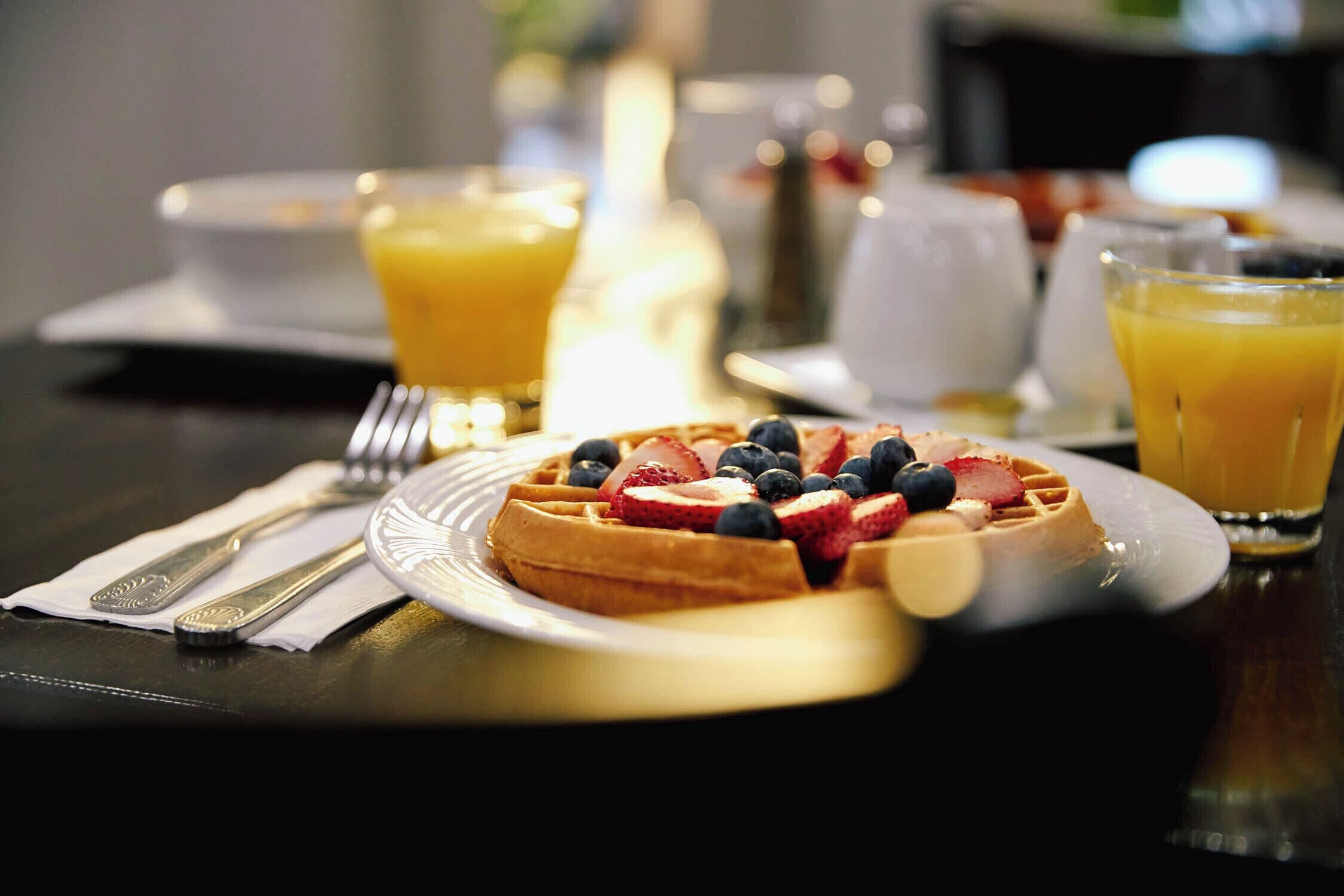 Ein Frühstückstisch mit einem Waffel, belegt mit Erdbeeren und Blaubeeren, einem Glas Orangensaft sowie einer Gabel und einem Messer auf einer Serviette.