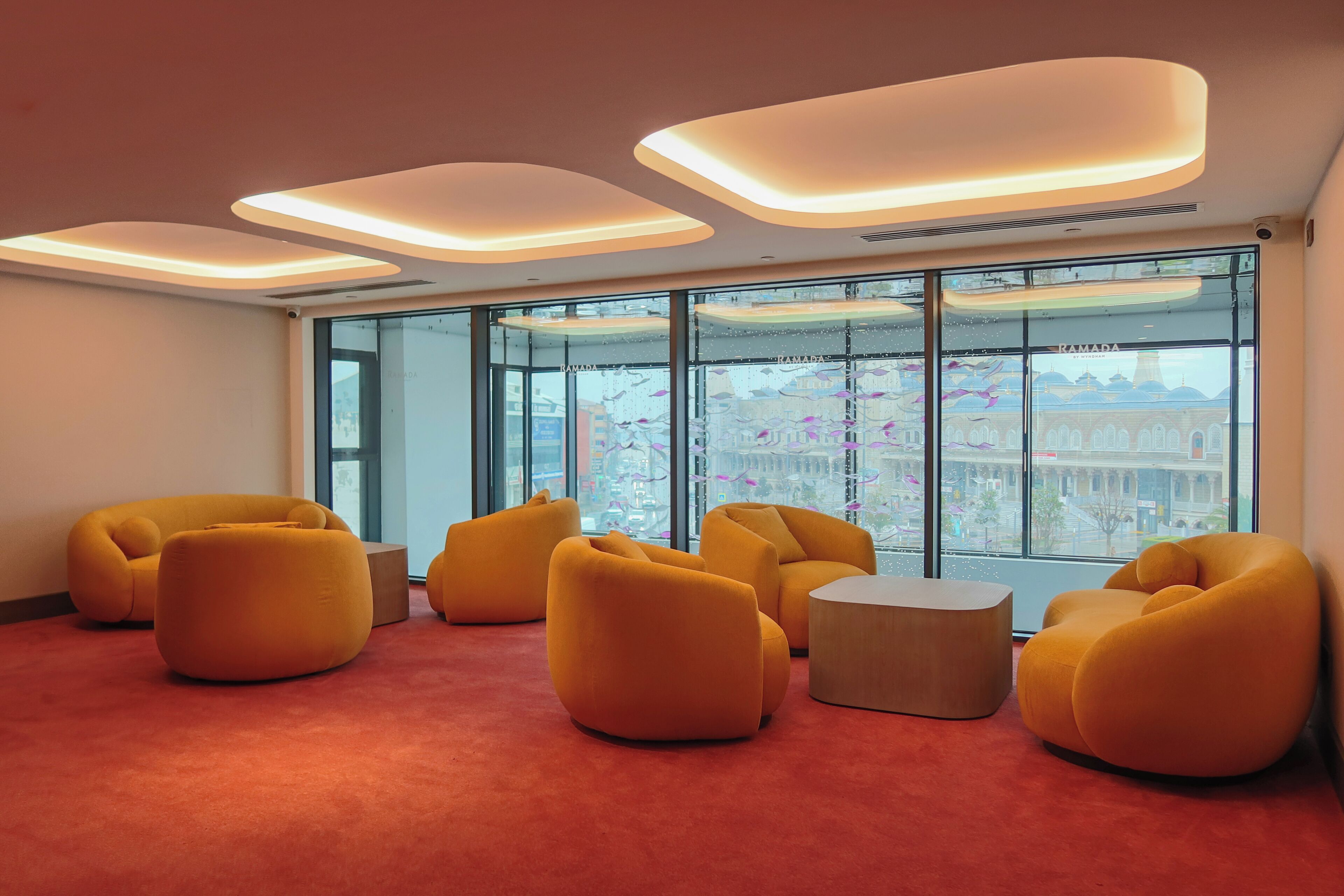 Un intérieur moderne avec des canapés orange et une moquette rouge, de grandes fenêtres offrant une vue sur la ville et des spots encastrés au plafond.