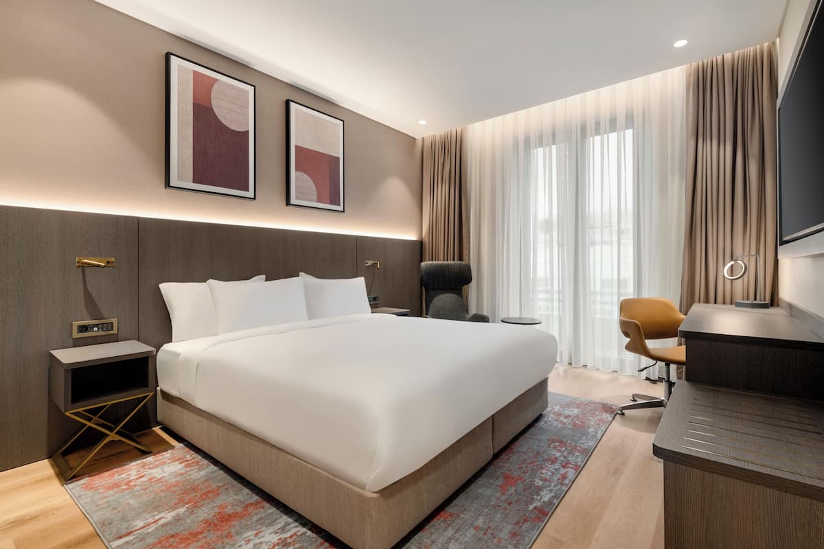 Habitación de hotel moderna con una cama grande, un escritorio y una silla. Hay dos cuadros en la pared, una mesita de noche y un televisor.