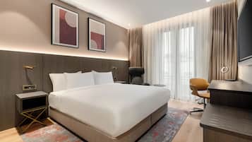 A modern hotel room with a large bed, a desk, and a chair. There are two framed pictures on the wall, a nightstand, and a TV.