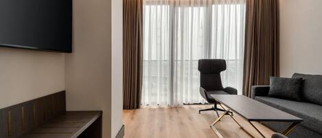 A modern hotel room with a flat-screen TV, a sofa, a dining table, and a large window with curtains.
