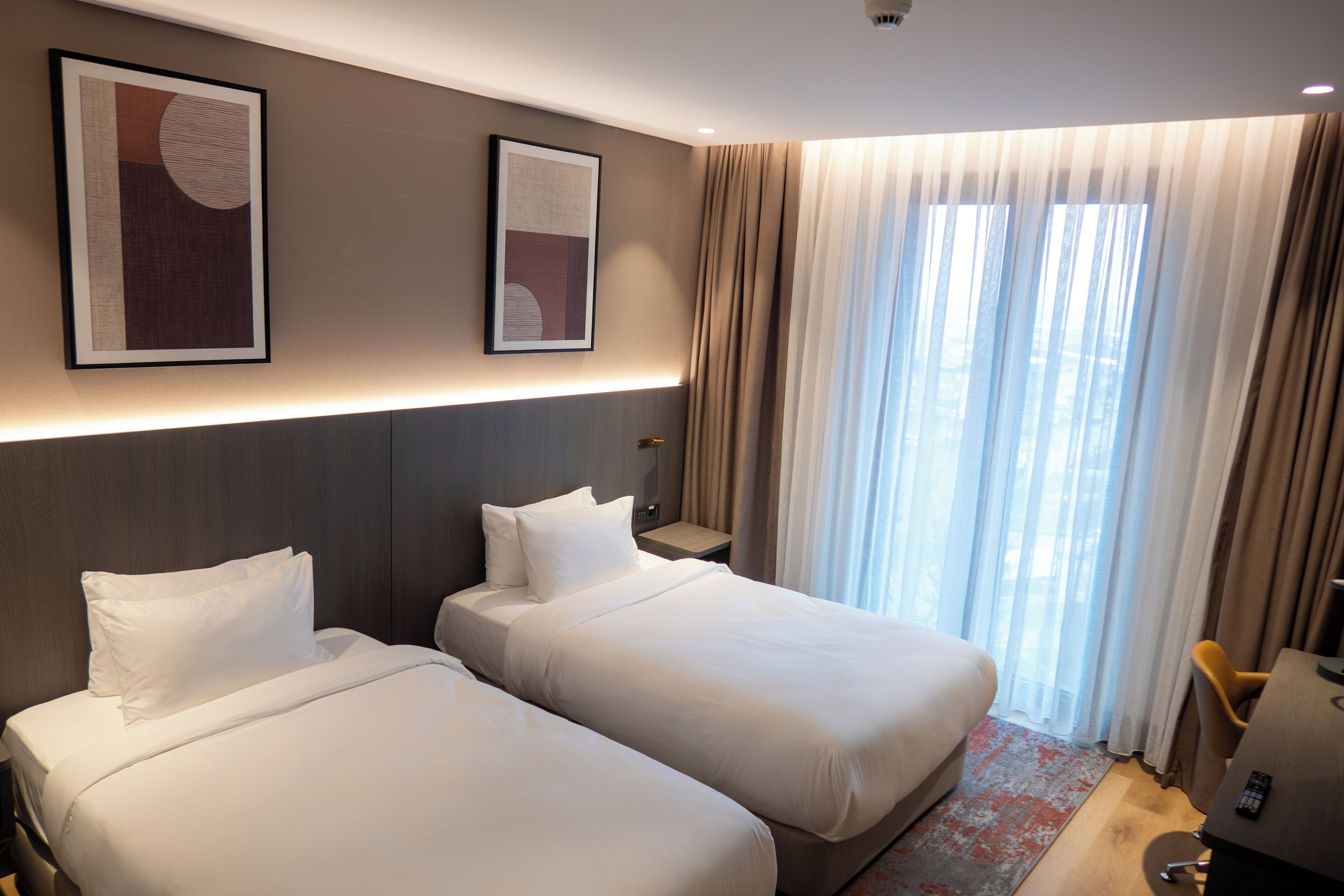 A hotel room with two beds, a desk, and a large window with curtains.
