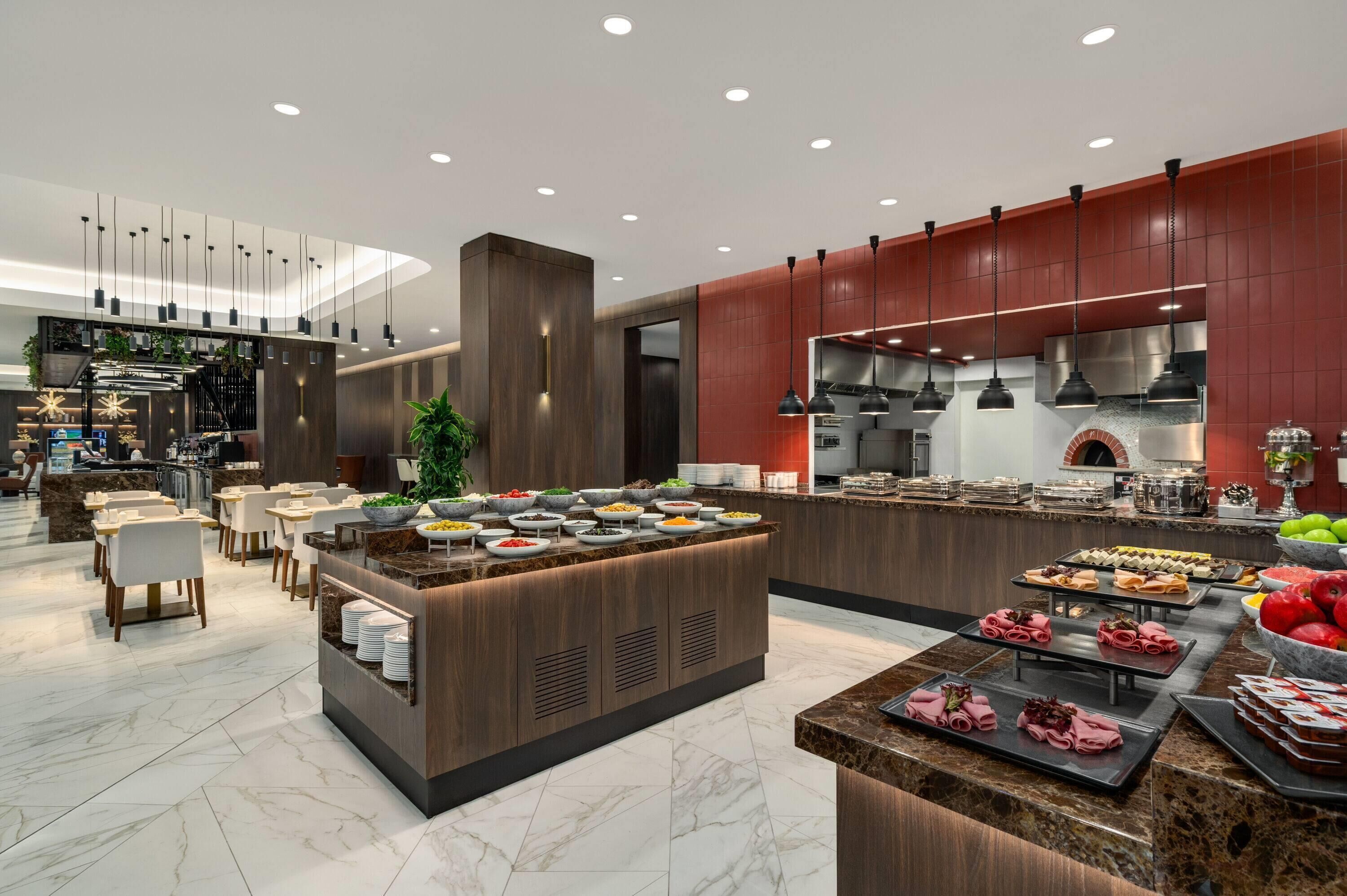 A modern hotel lobby with a central food station, seating area, and a visible kitchen.