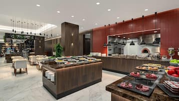 A modern hotel lobby with a central food station, seating area, and a visible kitchen.