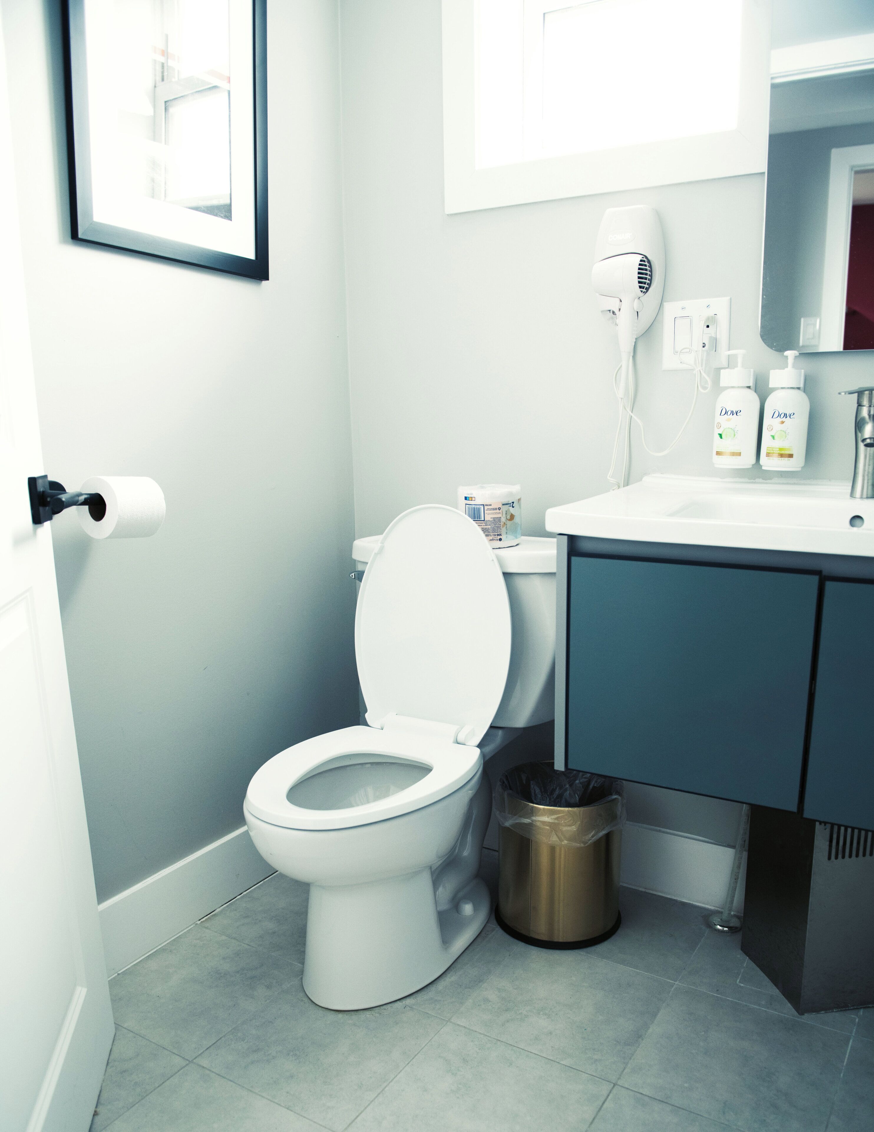 A bathroom with a toilet, a trash can, a hairdryer, and a sink with toiletries.