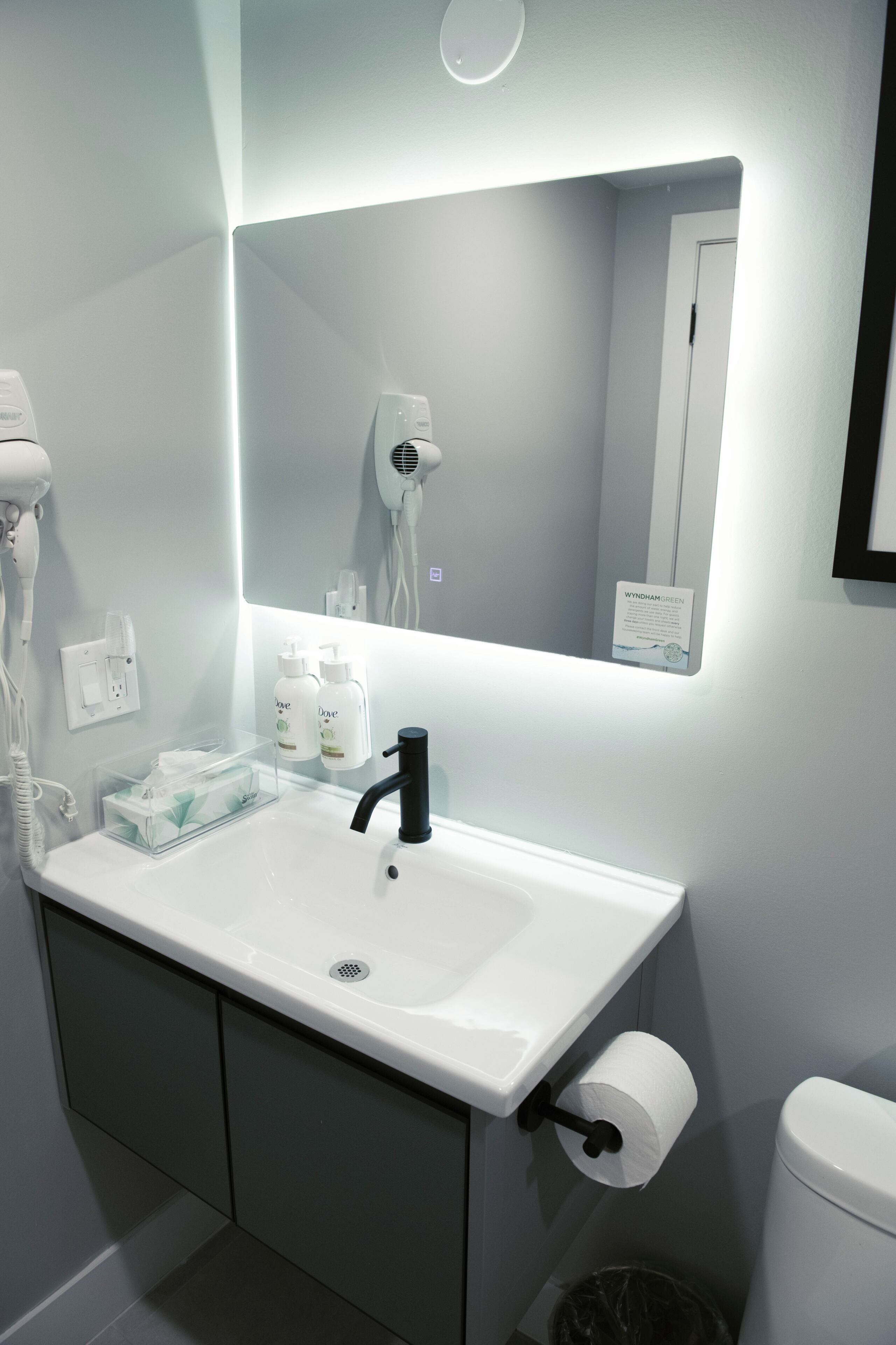 Un baño con lavamanos blanco, grifo negro y un espejo encima. Hay secadores de pelo y artículos de aseo en la pared.