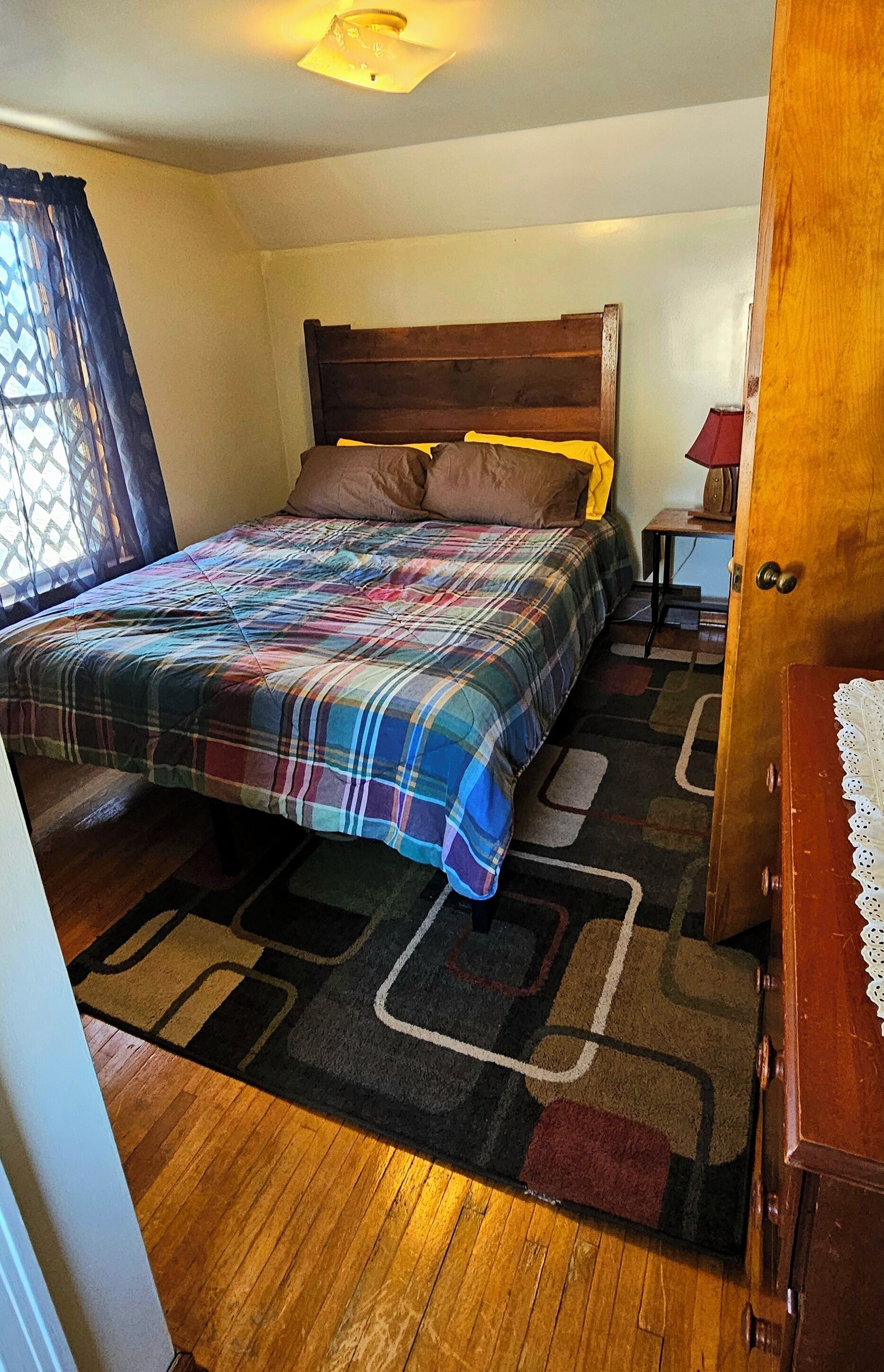A bedroom with a wooden bed, a colorful plaid bedspread, a small bedside table with a lamp, and a wooden nightstand.