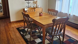 A dining area with a wooden table and chairs, a patterned rug, a hanging lamp, and a window with curtains. - Smitty's Cozy Corner in Smethport, PA. Spaciously 4-bedroom home (Smethport)