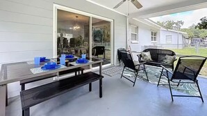 En täckt uteplats med ett bord och stolar, en takfläkt och utsikt över ett hus i bakgrunden.